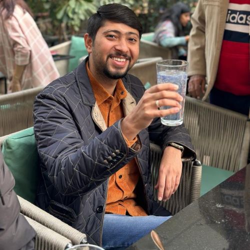 A cheerful person sits at a table outdoors, smiling and holding up a glass of water. They are wearing a light orange shirt under a dark quilted jacket. The setting appears to be a relaxed café with greenery in the background and other people in the distance.