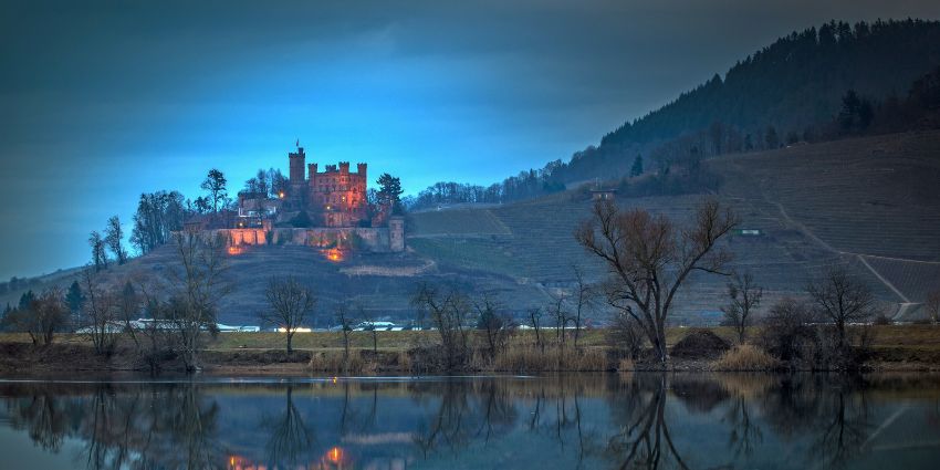 Photography Advice for a castle illuminated at dusk overlooks a calm lake, surrounded by trees and rolling hills, creating a tranquil landscape.