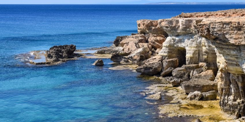 Picture-taking tricks for a Rocky cliffs rise beside clear turquoise waters, with scattered boulders and a tranquil sea extending to the horizon under a blue sky.