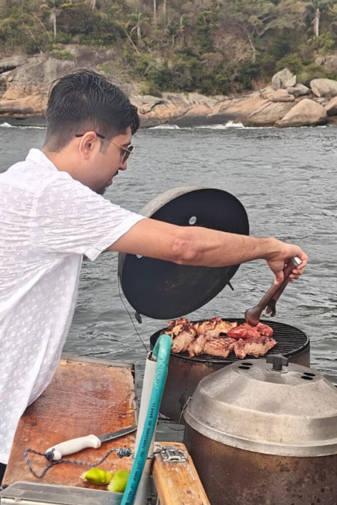 A person with dark hair and sunglasses is grilling meat on a portable grill while on a boat. The ocean and rocky shoreline are visible in the background. A wooden table is beside the grill, with a knife and two limes placed on it.